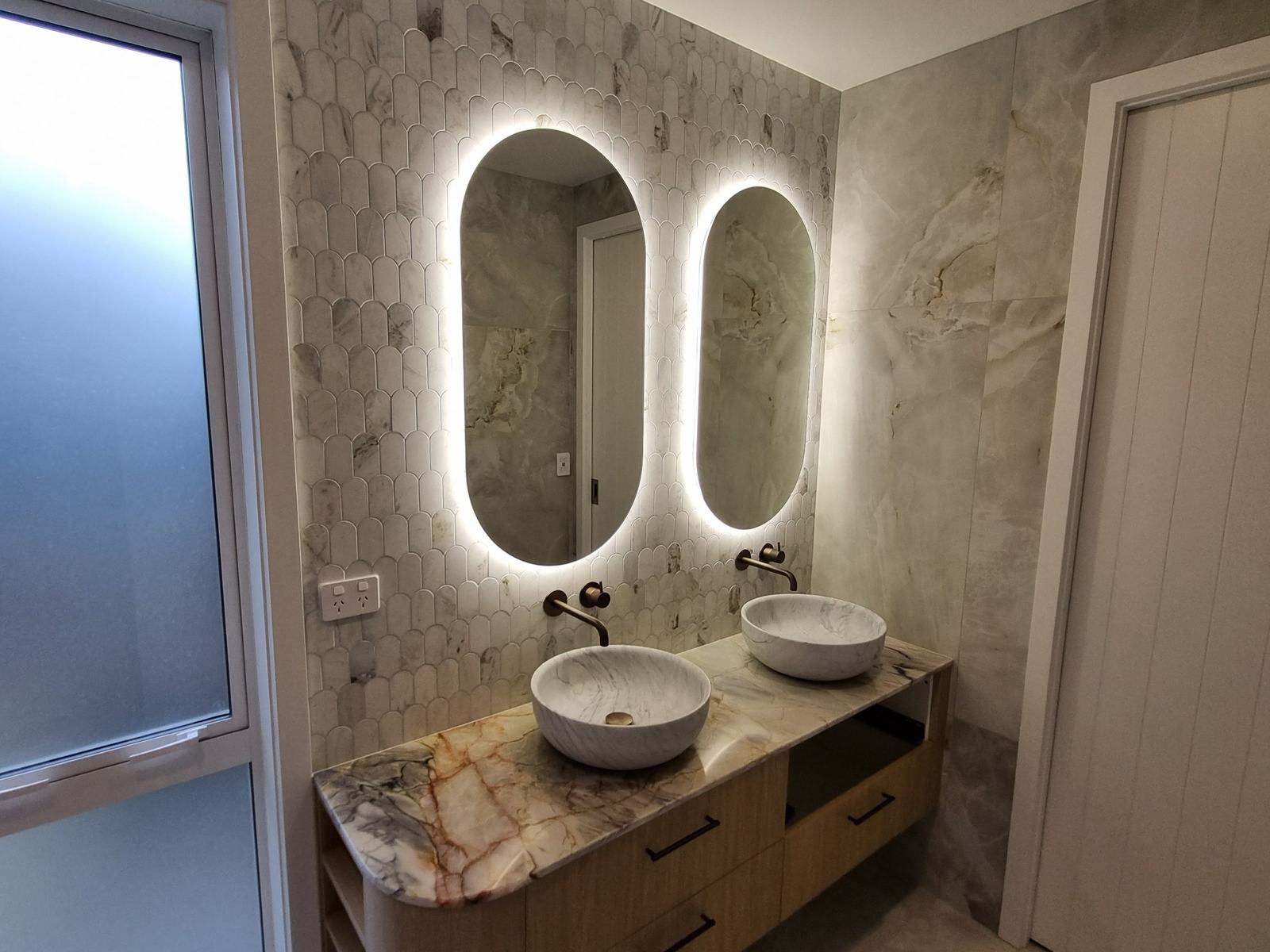 Marble double vanity with backlit oval mirrors and fish-scale tile wall