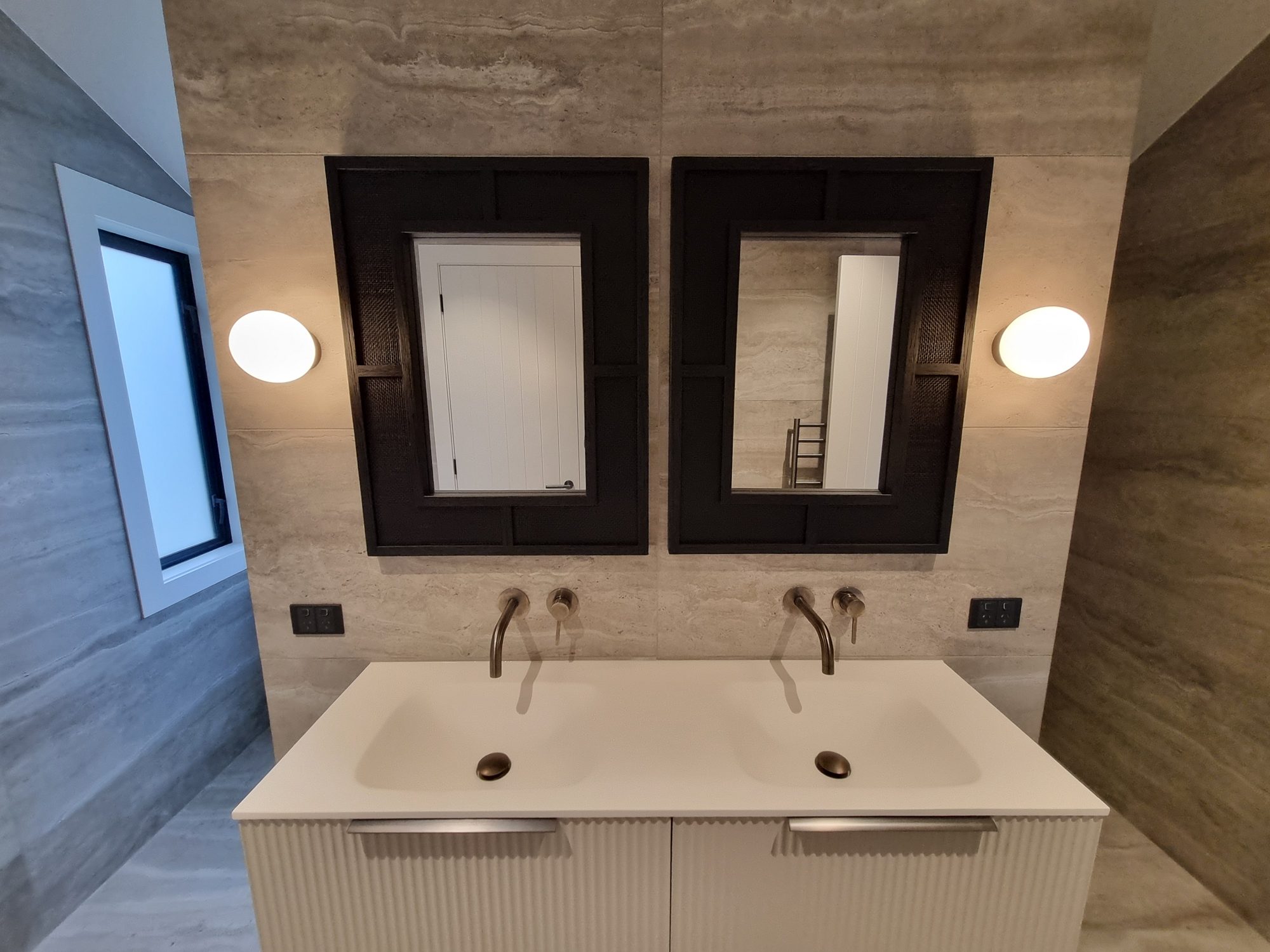 Floating fluted double vanity with two black framed mirrors, round wall lights and wall-mounted taps against large-format tile