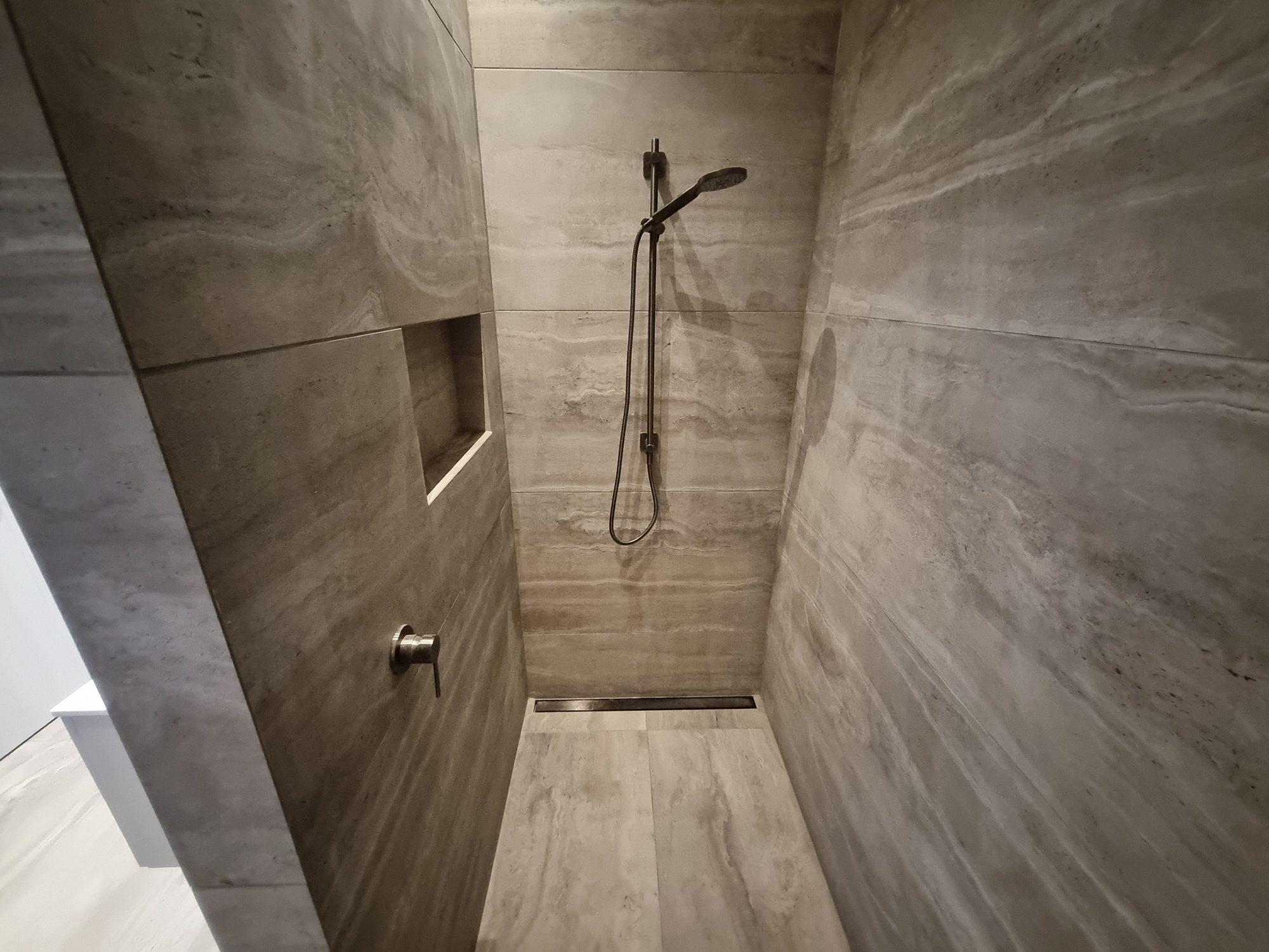 Walk-in shower fully lined in large-format travertine-look tile with a recessed niche, slide rail and linear drain