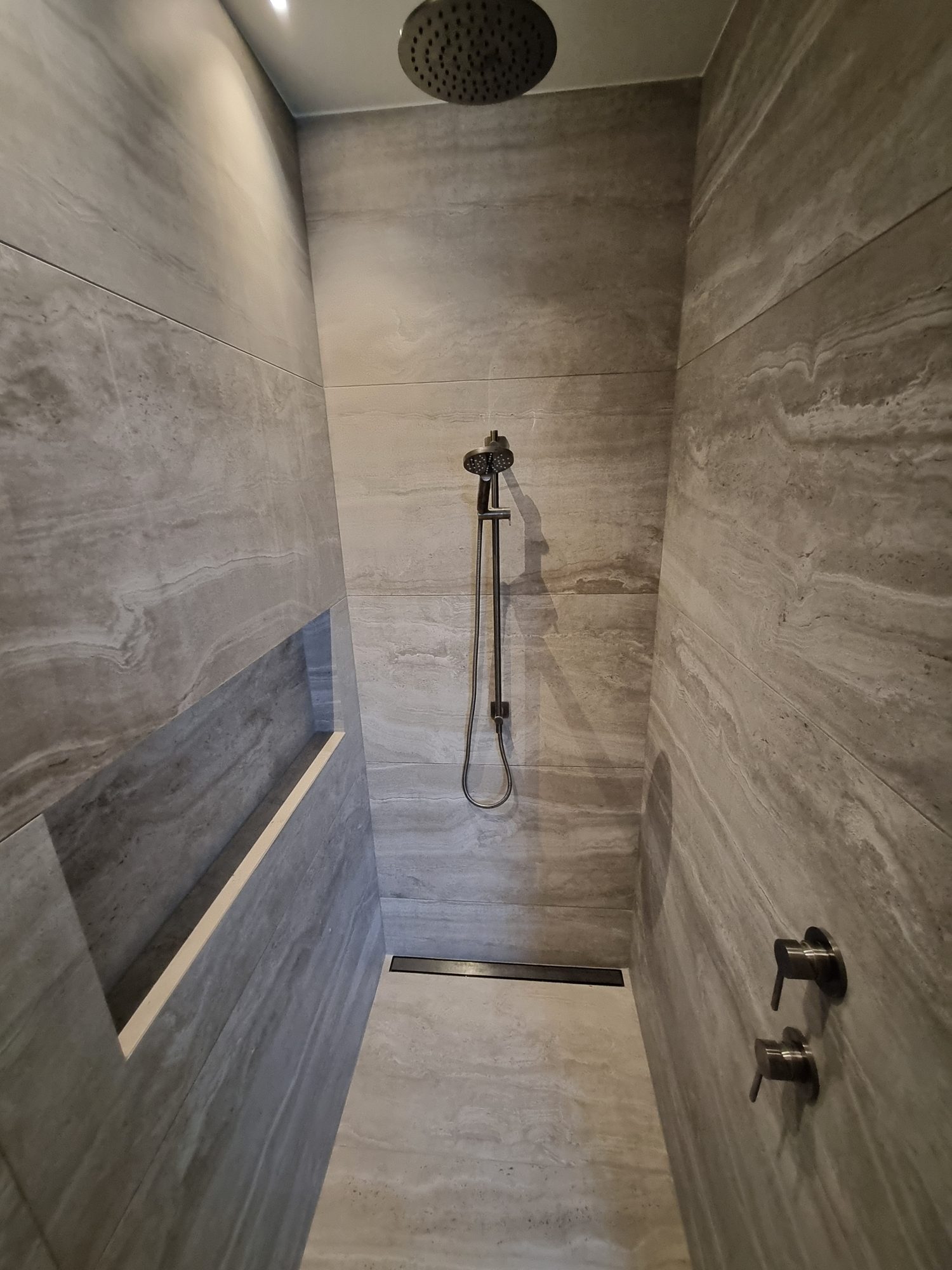 Walk-in shower fully lined in large-format travertine-look tile, with a rain head, slide rail, linear drain and a tiled niche