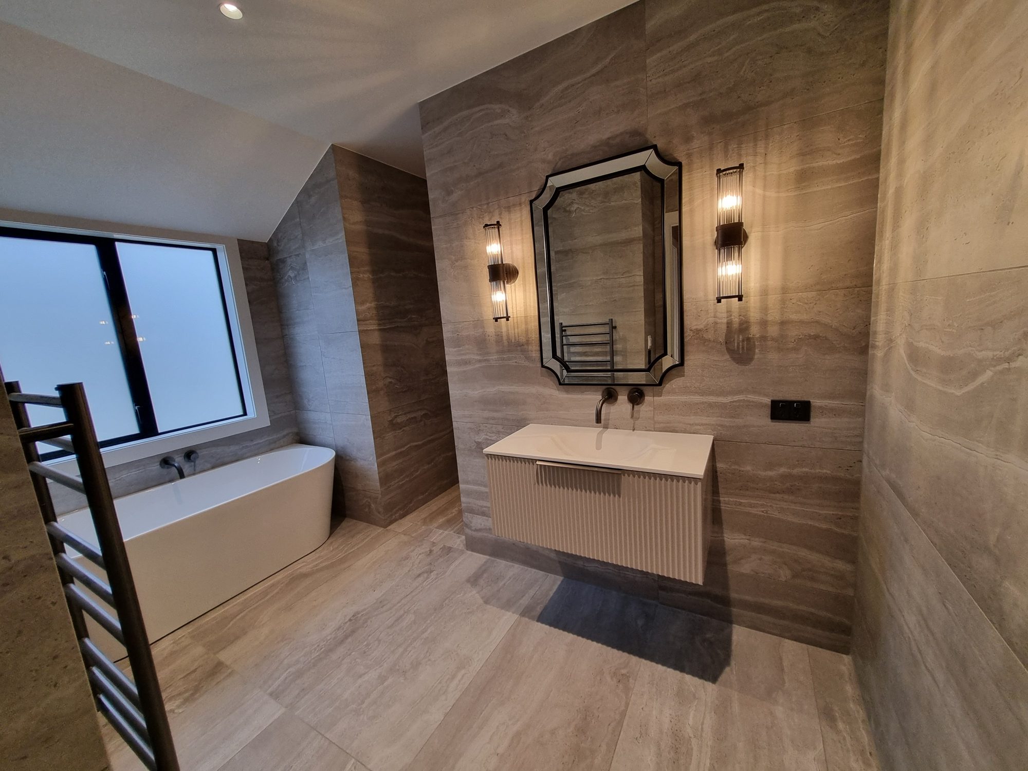 Palmer Mill guest ensuite with a freestanding bath under the window, a floating fluted vanity and shaped mirror, all in large-format travertine-look tile