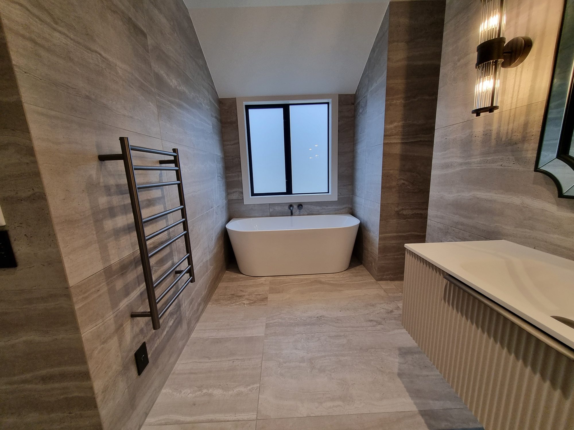 Freestanding bath set against large-format travertine-look tile under a window, with a heated towel rail and the floating vanity in the foreground