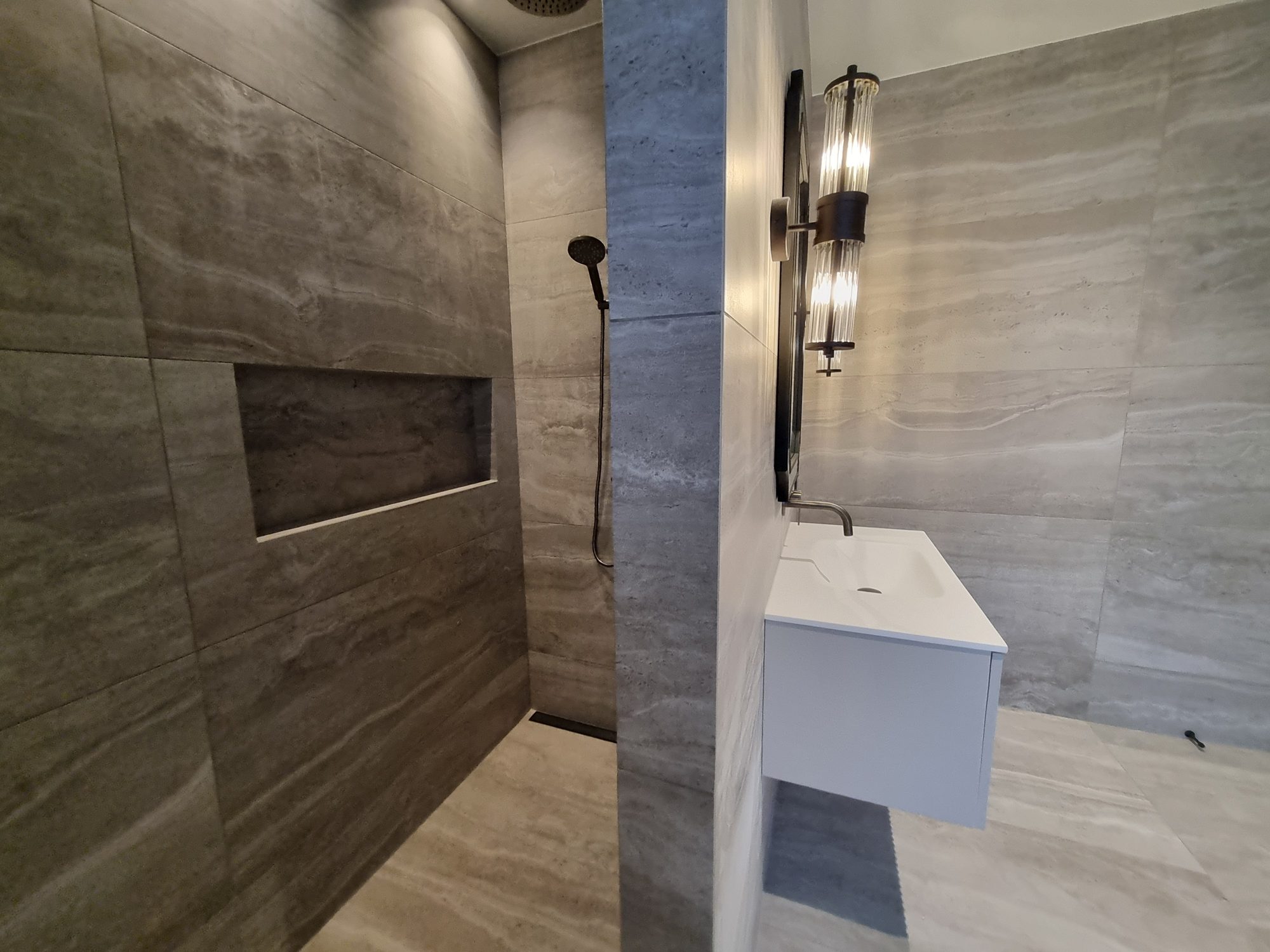 Palmer Mill guest ensuite shower with a recessed tiled niche in large-format travertine-look tile, beside a floating vanity and glass wall light