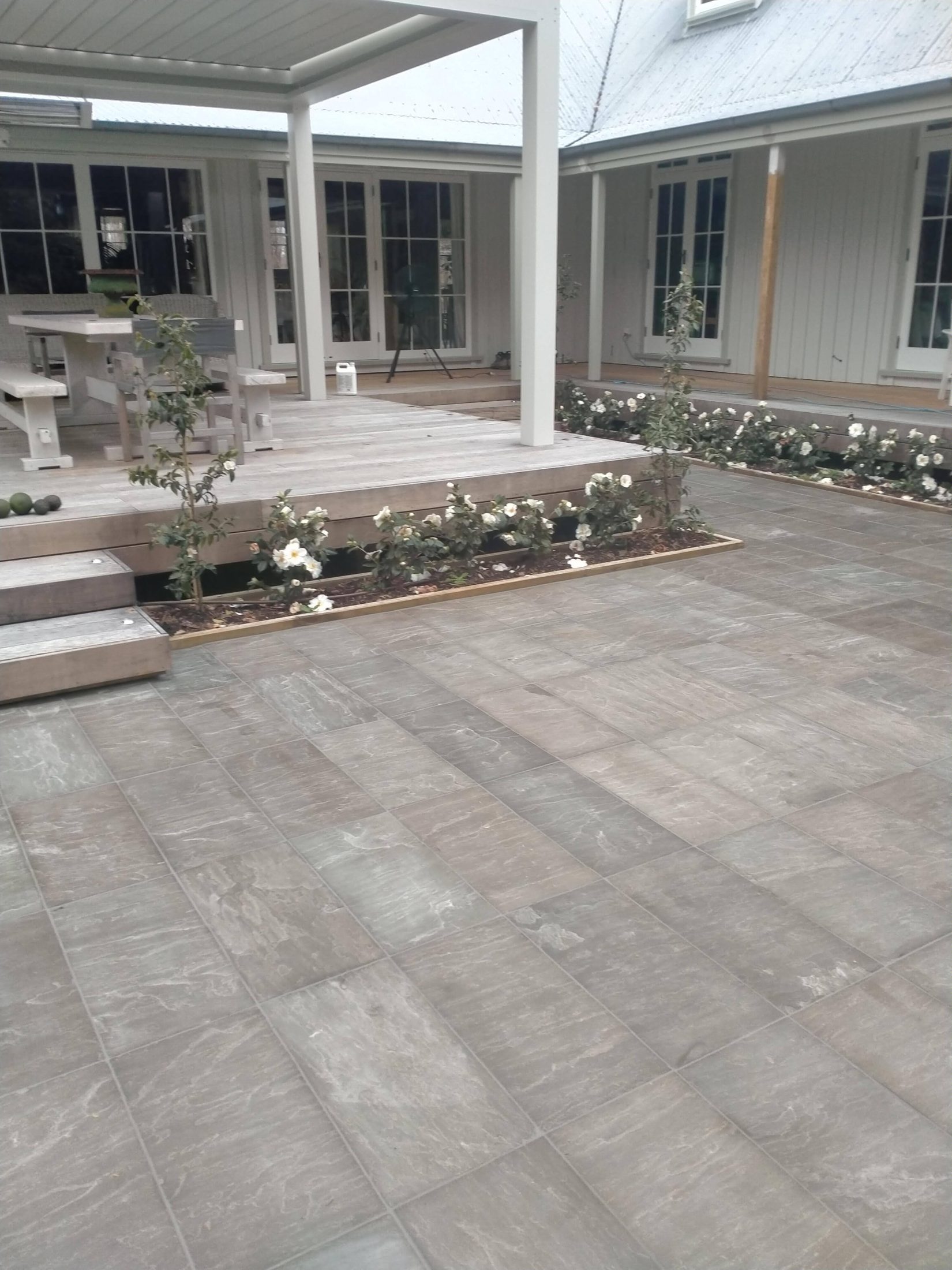 Large-format sandstone paving in a covered courtyard beside a deck with steps and camellias, Gisborne