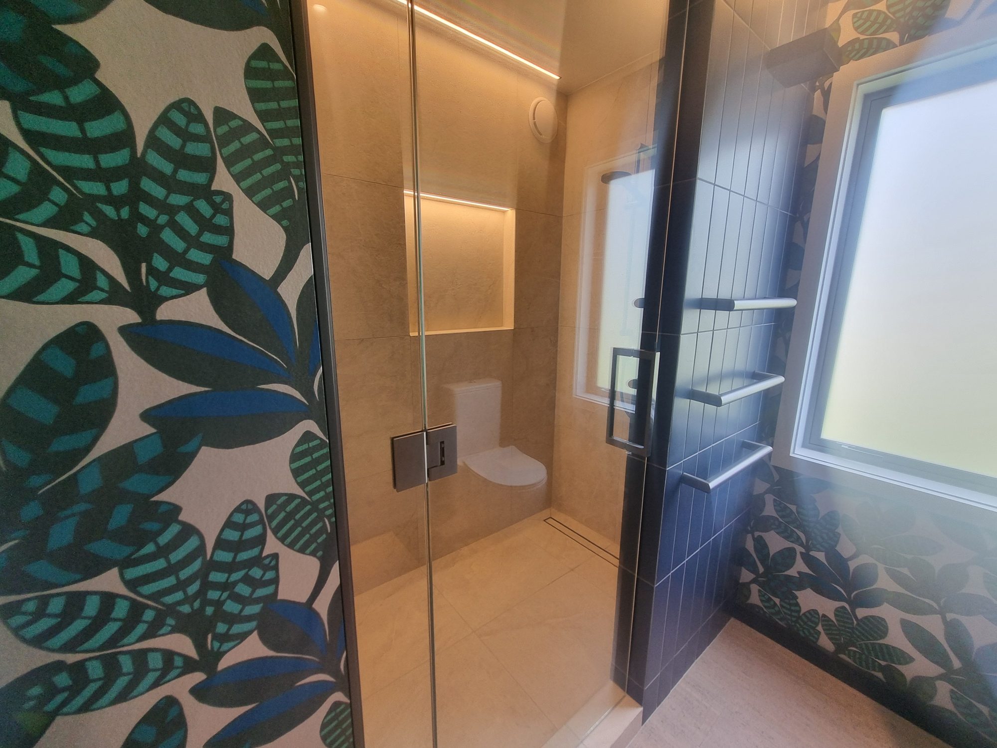 Glass shower screen and large-format tiled shower with lit niche, beside a leaf-pattern wallpapered wall and dark vertical tiles