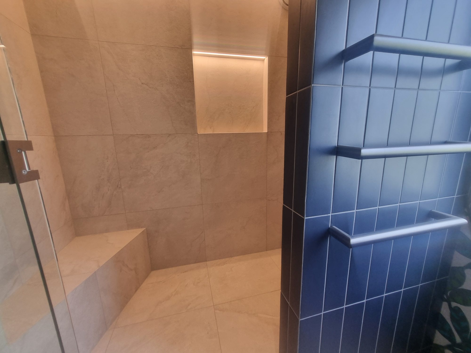 Large-format tiled shower with a tiled corner bench, lit niche and linear drain, beside blue vertical tiles with towel rails