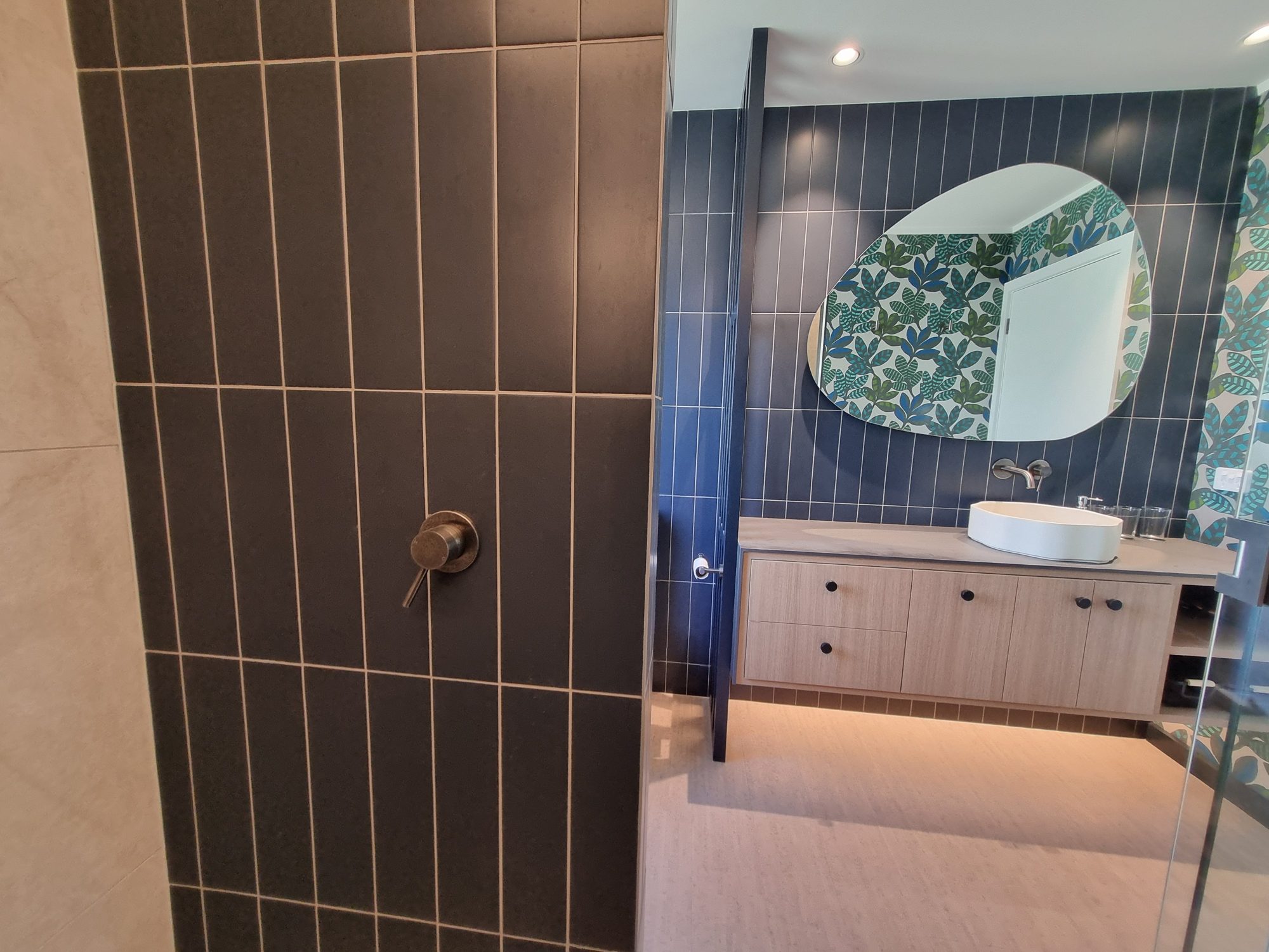 Dark vertical-stack tiled shower wall with a brushed mixer, looking across to the floating vanity and organic-shaped mirror