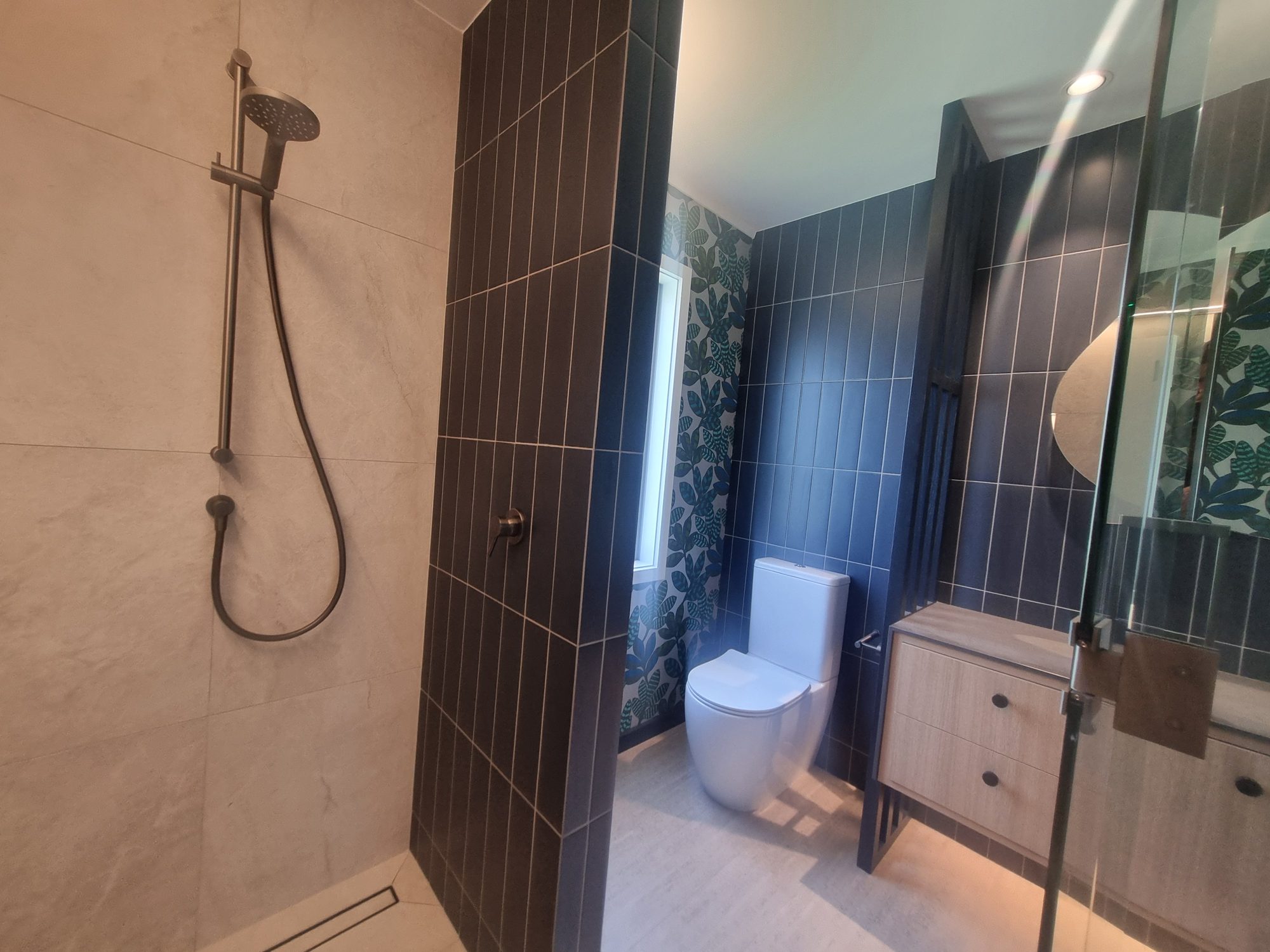 Waitetoko bathroom walk-in shower with large-format light tiles, chrome shower rail, dark vertical-stack partition wall and linear drain
