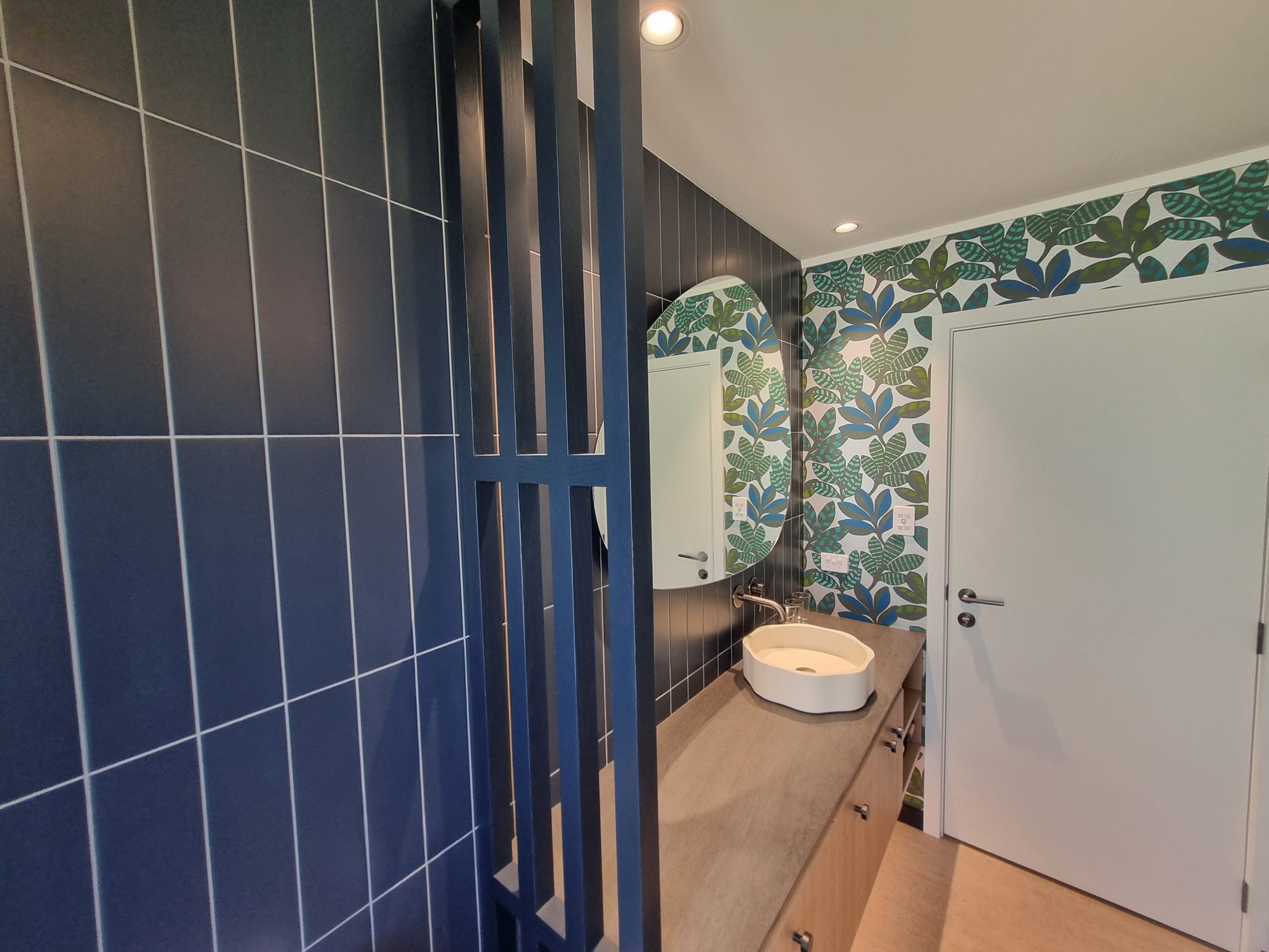 Waitetoko bathroom vanity beyond a dark timber slat screen, with navy vertical tiles, organic-shaped mirror and leaf-pattern wallpaper