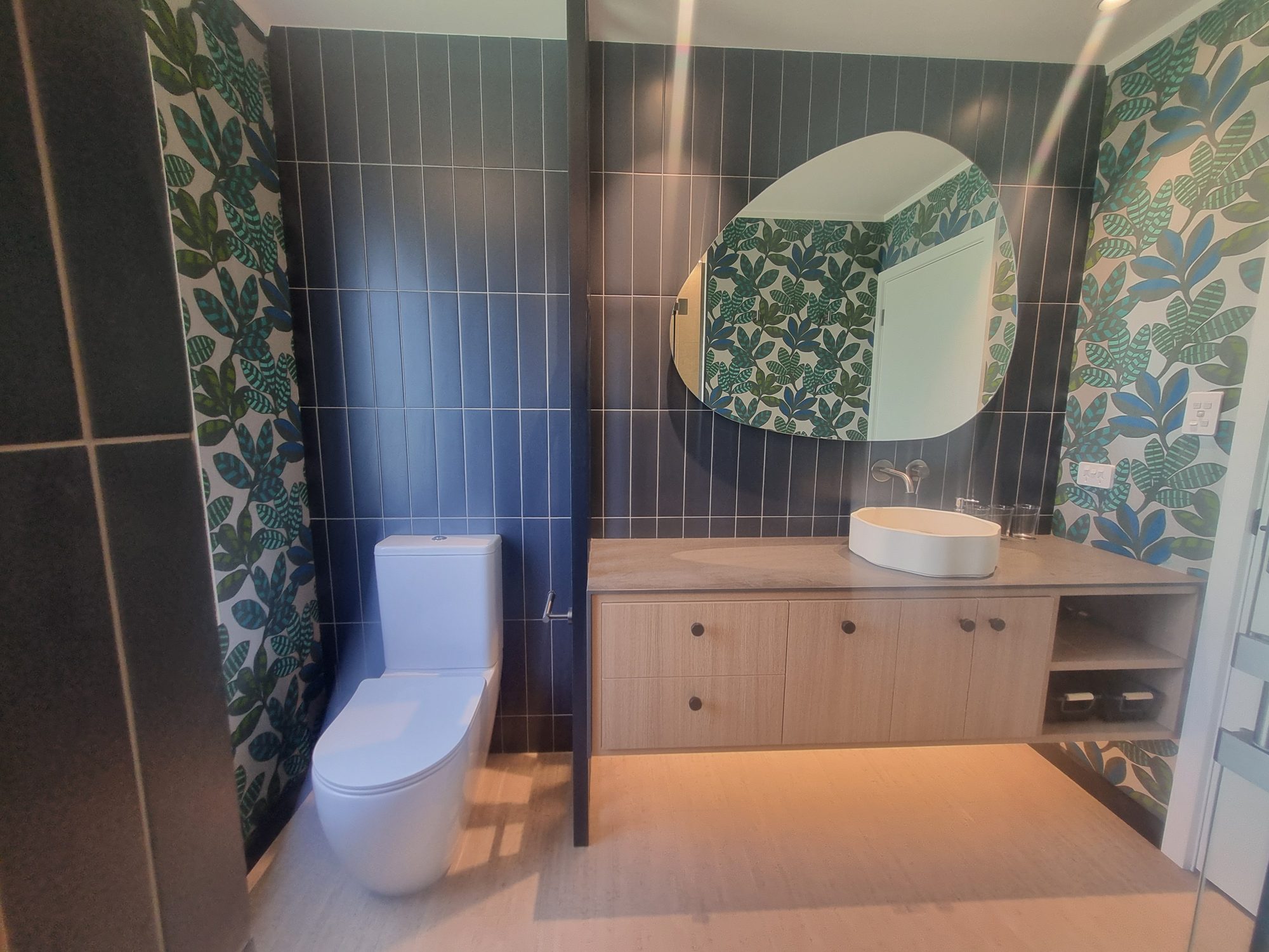 Waitetoko bathroom vanity with dark navy vertical-stack tiles, organic-shaped mirror, floating timber vanity and leaf-pattern wallpaper