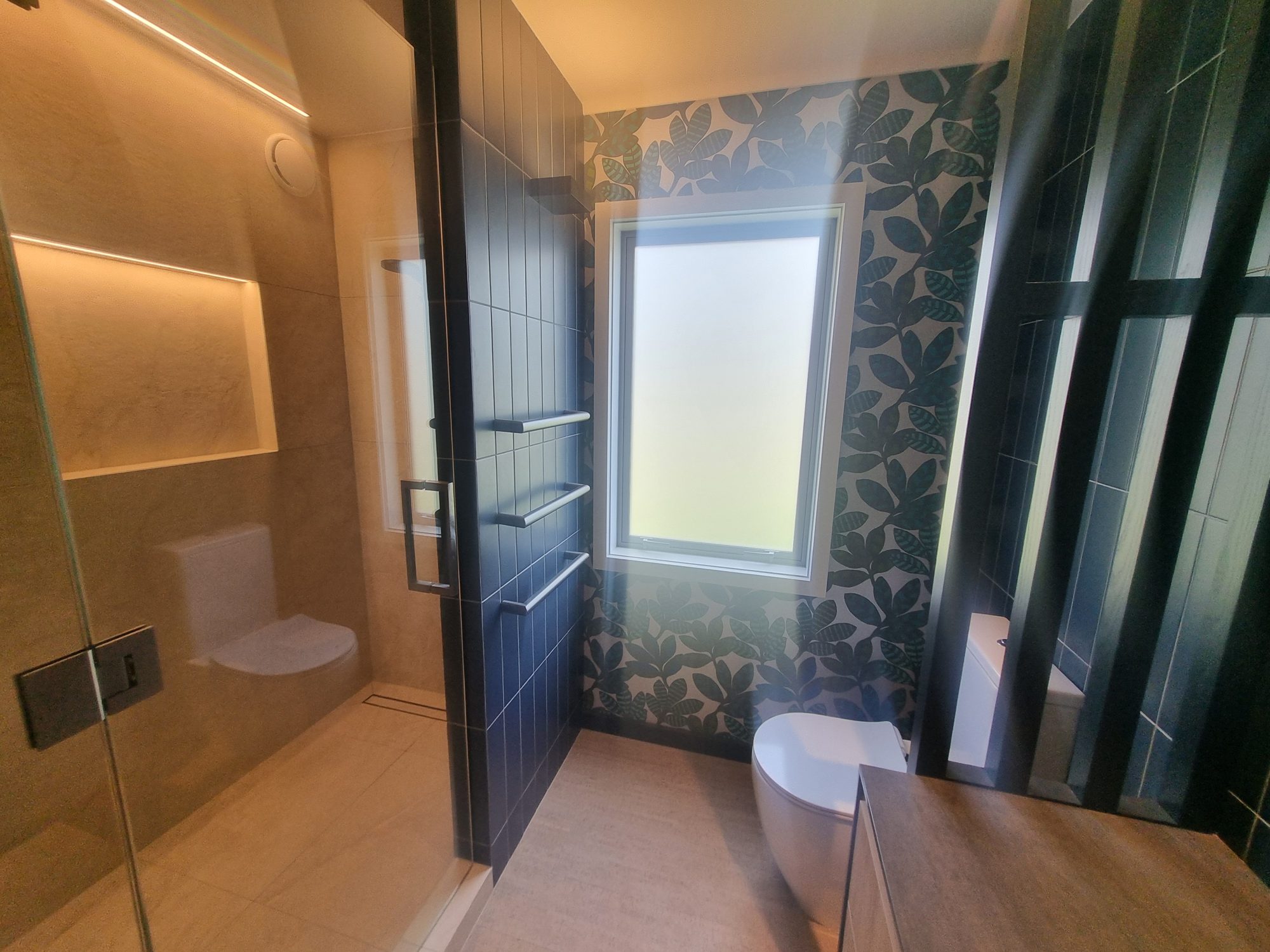Waitetoko bathroom shower seen through the glass door, with a lit niche, linear drain, towel rails and leaf-pattern wallpaper