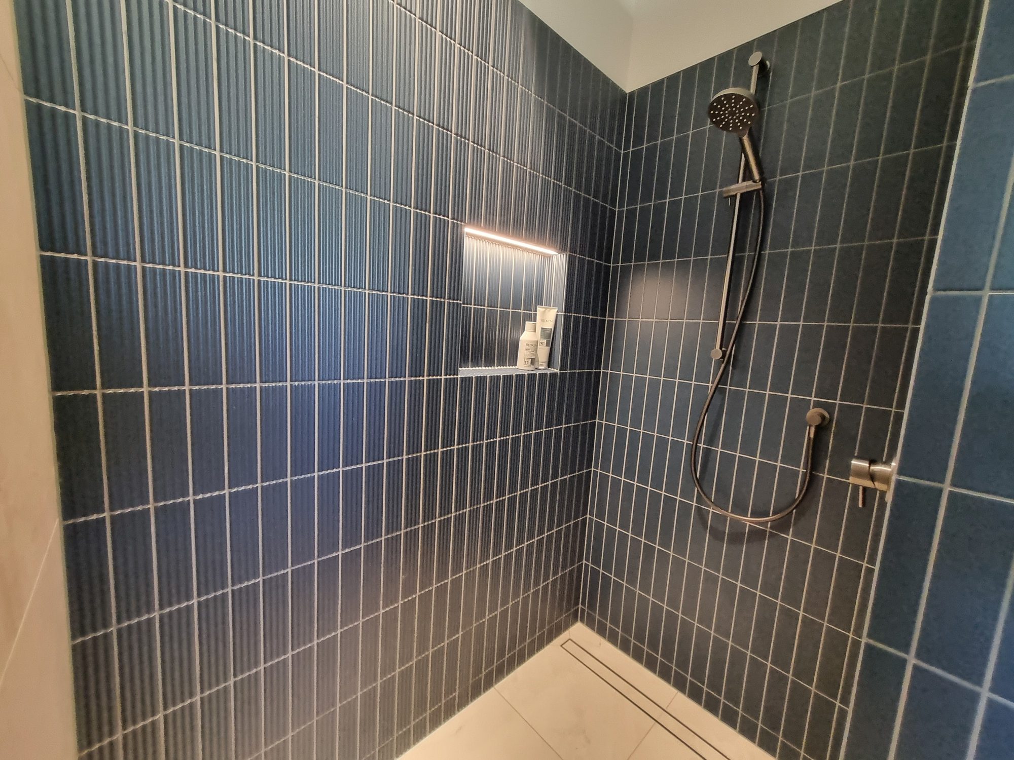 Walk-in shower lined with fluted dark blue tiles, a lit recessed niche, chrome shower rail and a linear drain over large-format floor tiles