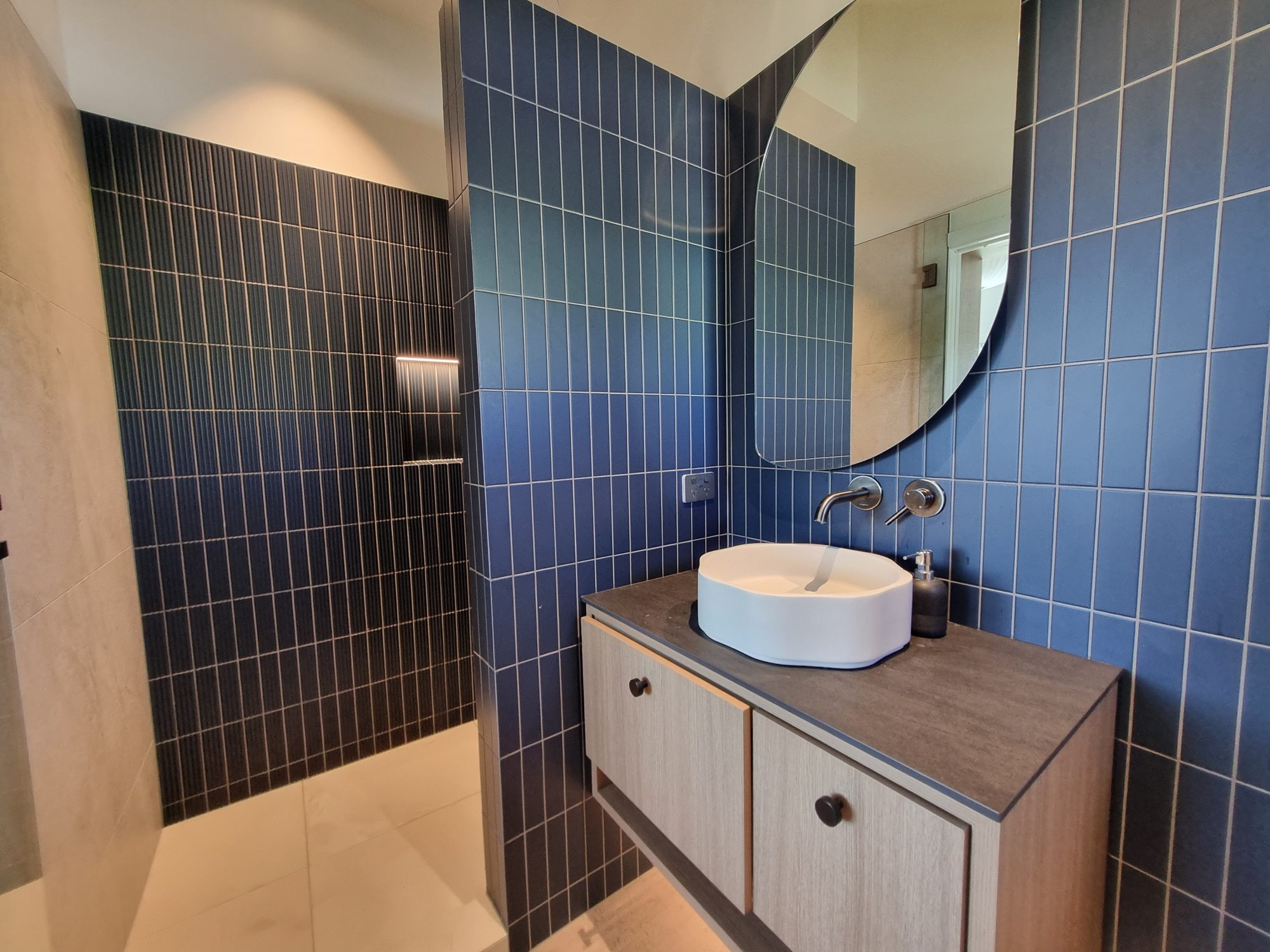 Ensuite vanity with a round mirror and wall-mounted taps against blue vertical-stack tiles, with the fluted blue shower wall beyond