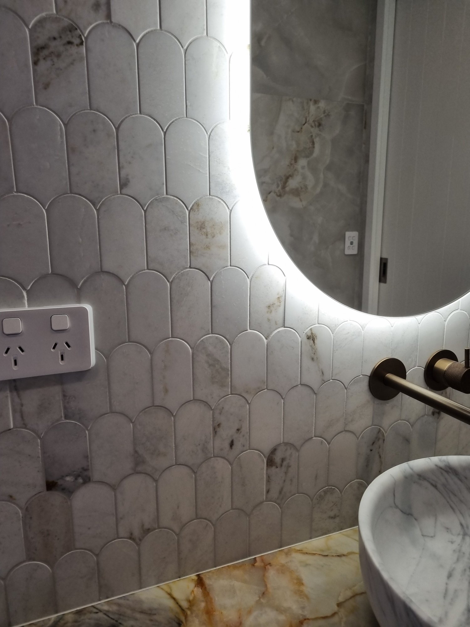 Close-up of marble fish-scale mosaic tiles with crisp grout lines, cut cleanly around a power point and a backlit oval mirror, with bronze tap and marble basin