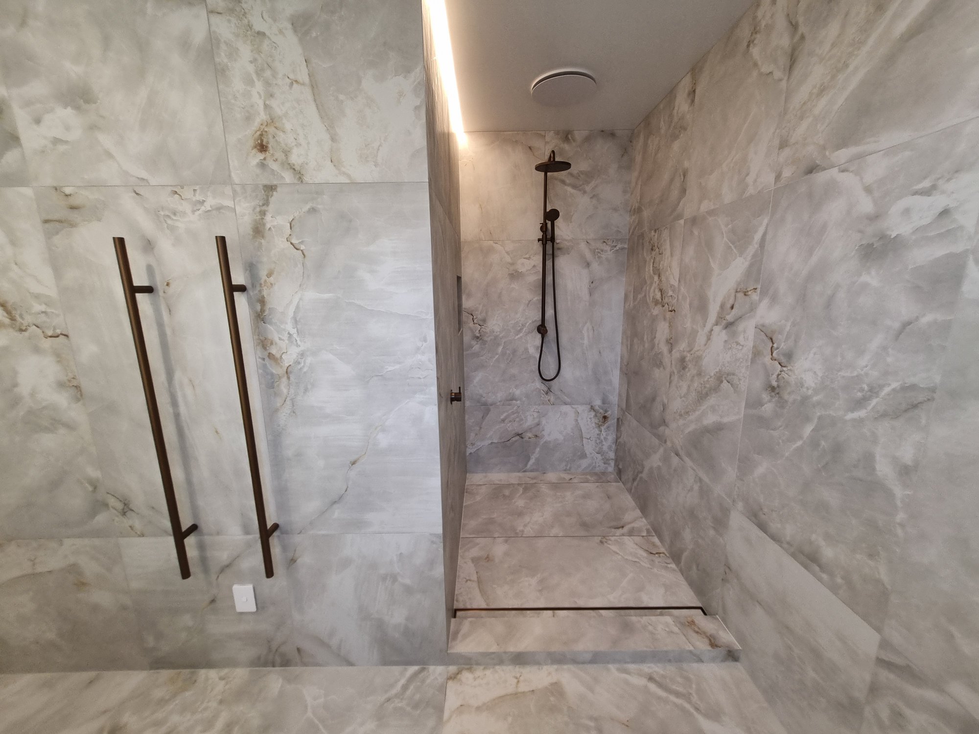 Walk-in shower lined with large-format onyx-look porcelain, with a tiled hob step, linear channel drain, shower niche and bronze shower fittings