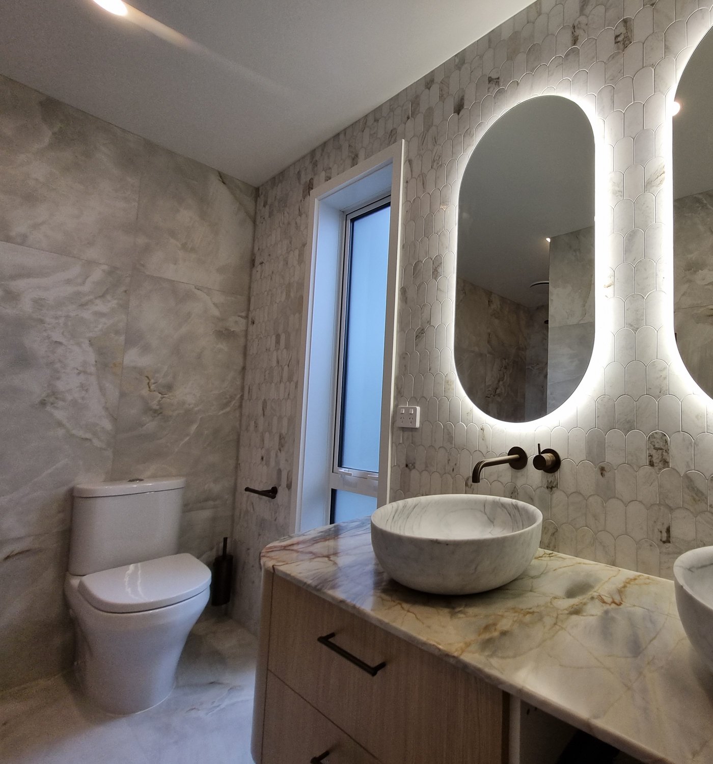 Ensuite vanity with a marble vessel basin, bronze tapware and a backlit oval mirror against a marble mosaic wall, beside large-format porcelain tile