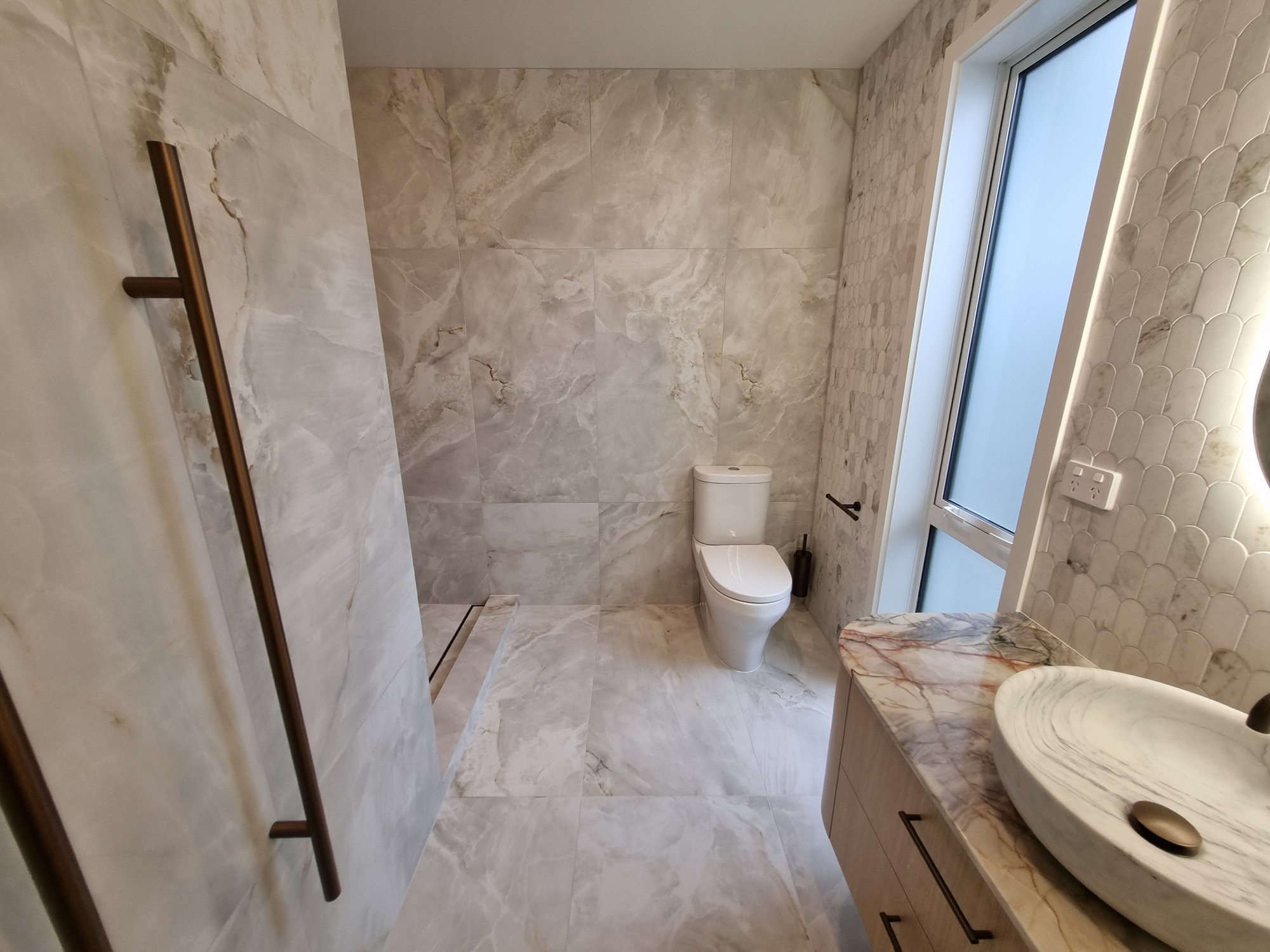 Wide view of a Taupō ensuite with large-format onyx-look porcelain walls and floor, a walk-in shower with hob step, toilet and vanity with marble mosaic wall